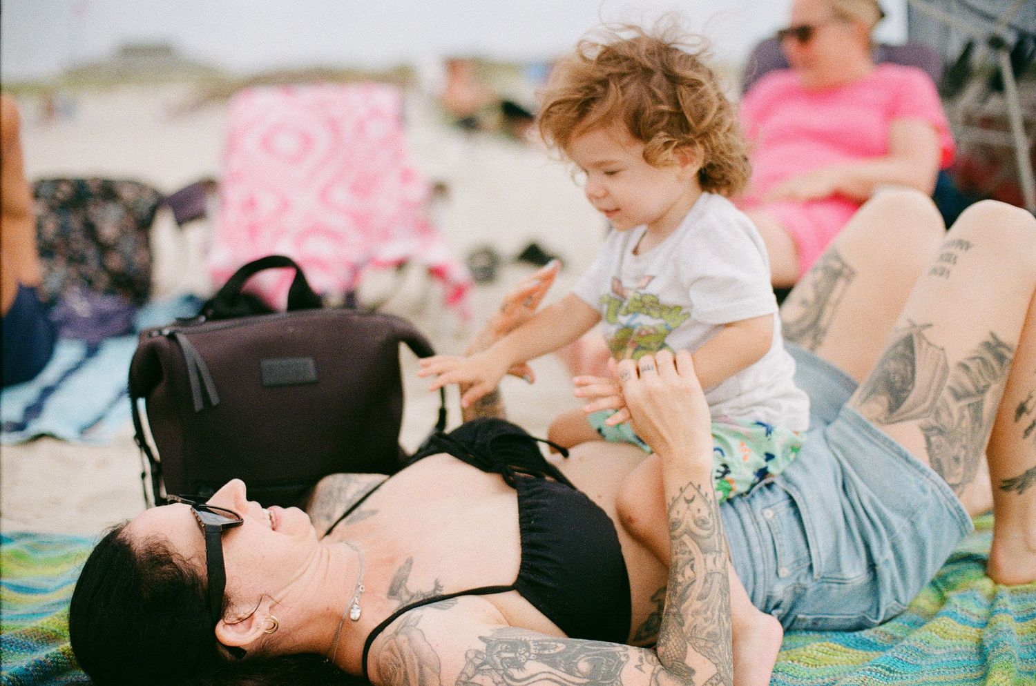 Tattooed mother lying on beach blanket playfully lifting smiling toddler in white shirt into the air on sunny day.