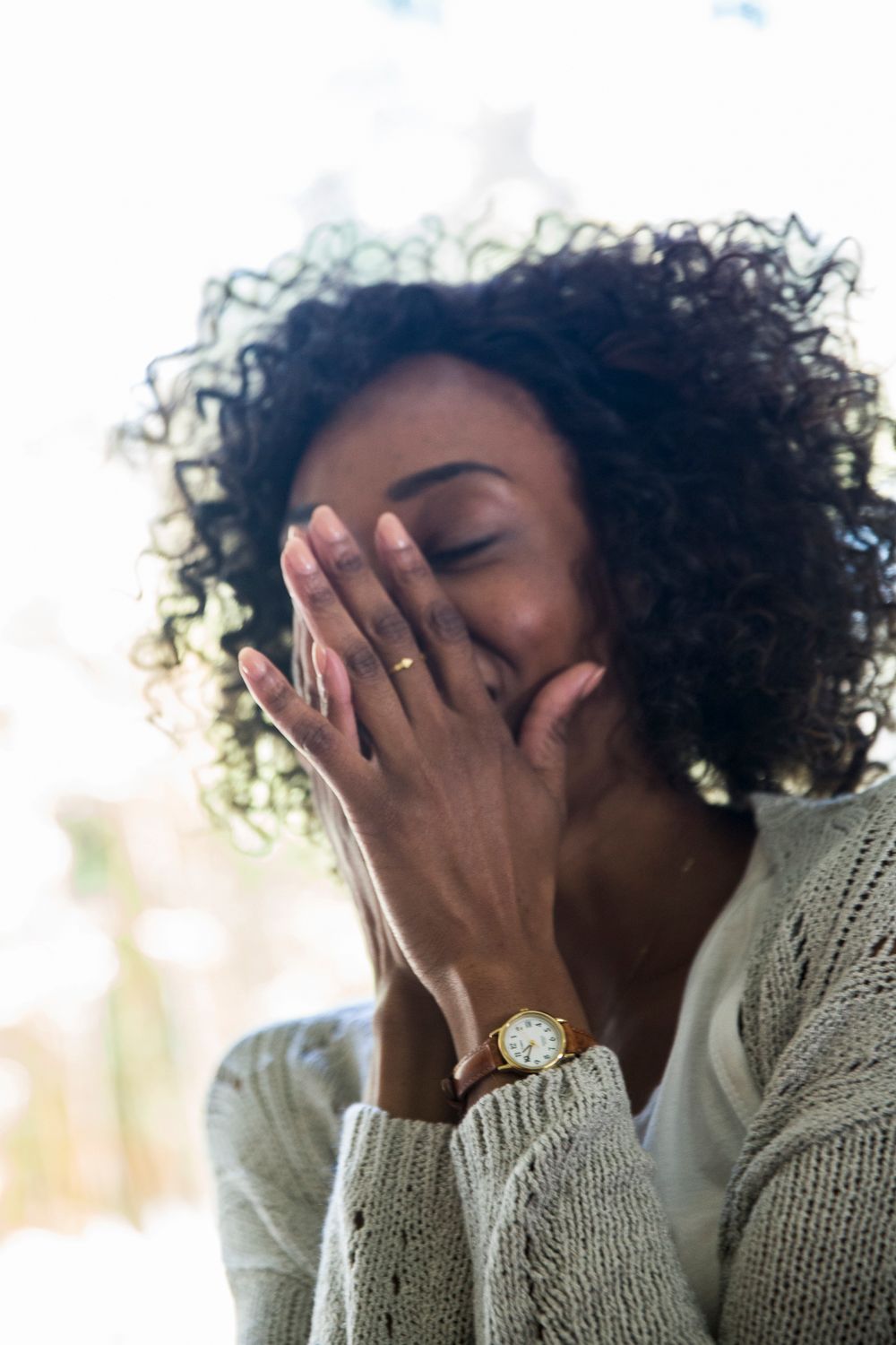 A young woman with curly hair laughs while covering her mouth with her hand, wearing a watch and ring.