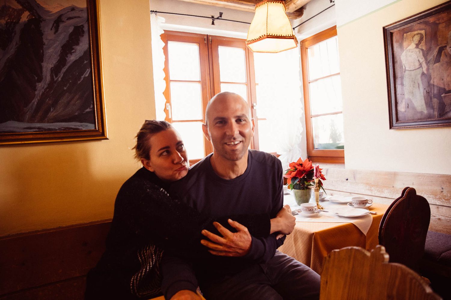 Couple embracing and smiling together at a cozy restaurant table with paintings and windows in background.