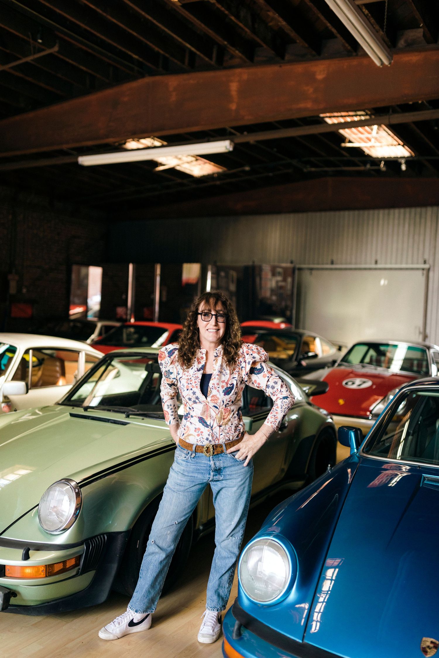 A woman poses confidently among classic Porsche cars in a garage, wearing jeans and a floral shirt.