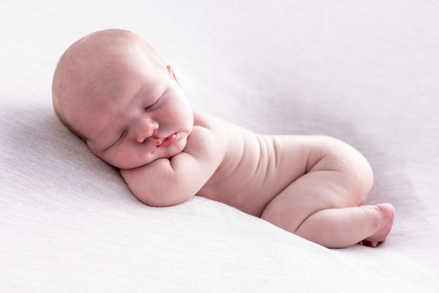 A sleeping newborn baby curled up in a peaceful pose on a soft white blanket. Poole Photographer