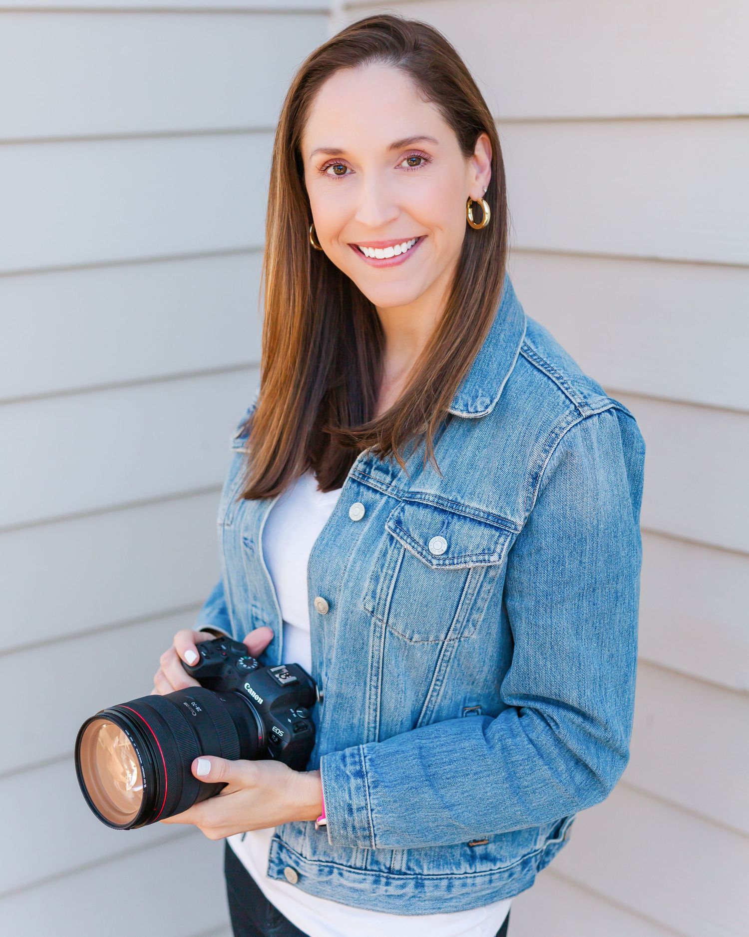 Professional photographer in denim jacket holding DSLR camera while posing against gray wall.