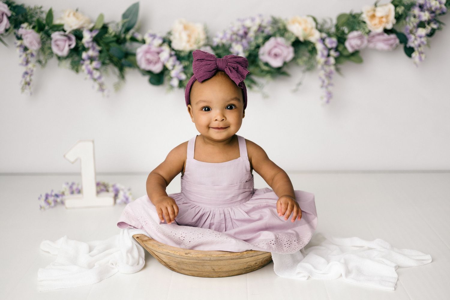 A child in a pink dress sits in a wooden bowl for their first birthday photoshoot with floral decor overhead.