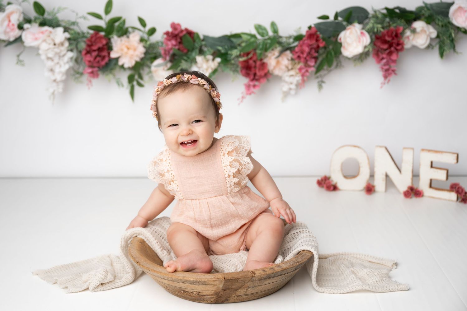 A small child in a pink romper sits in a wooden bowl against a white backdrop with floral decorations and 'ONE' letters.