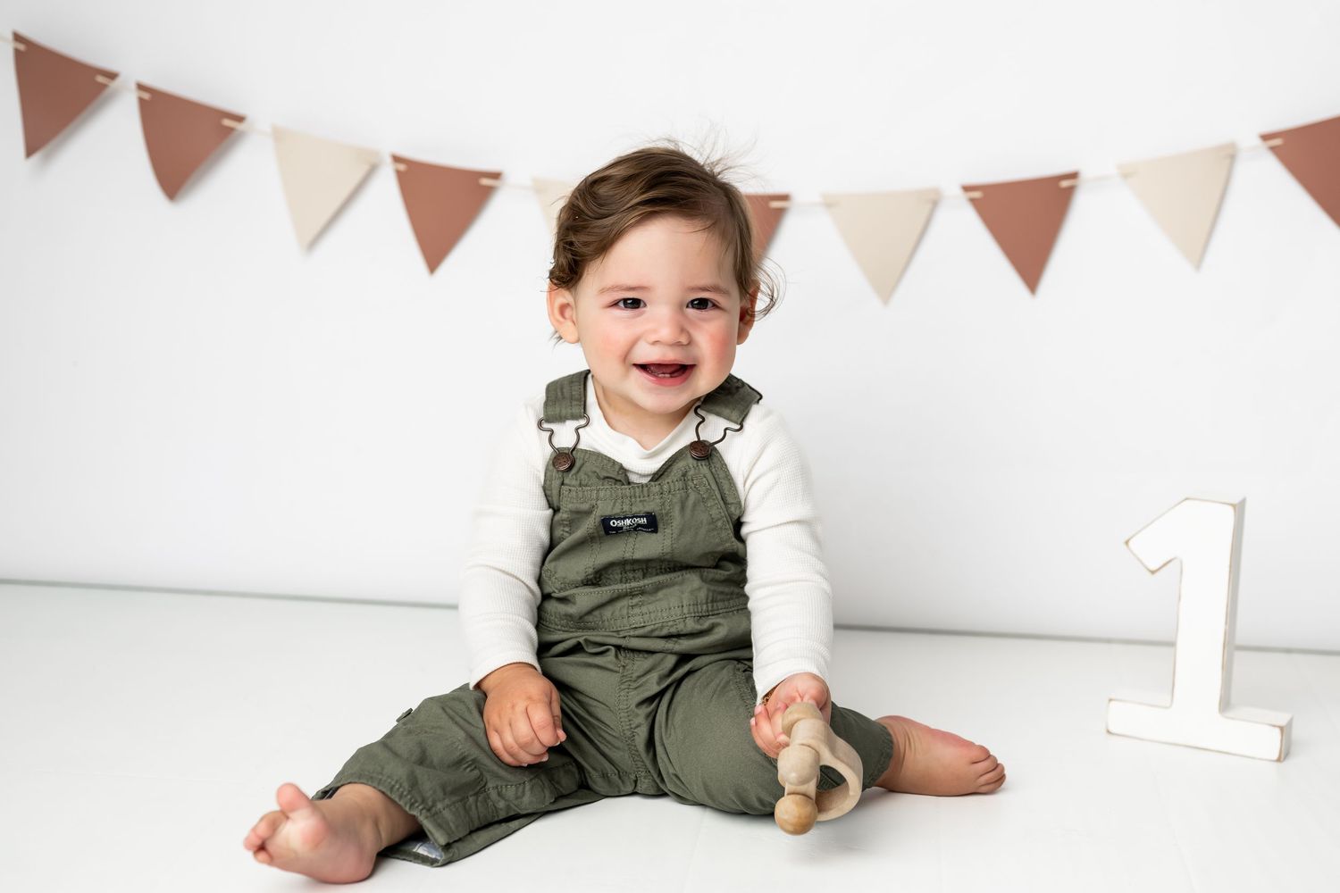 A small child wearing olive green overalls and white shirt sits on floor with festive triangle banner decorations above.
