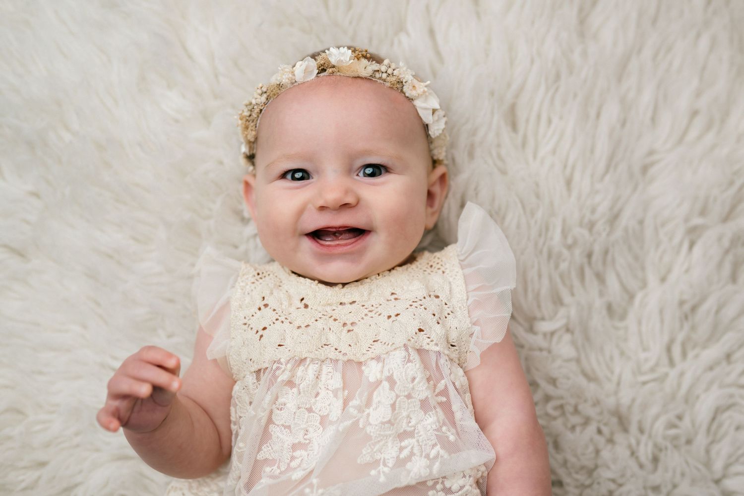 Smiling baby wearing white lace dress and floral headband lying on soft white fur background.