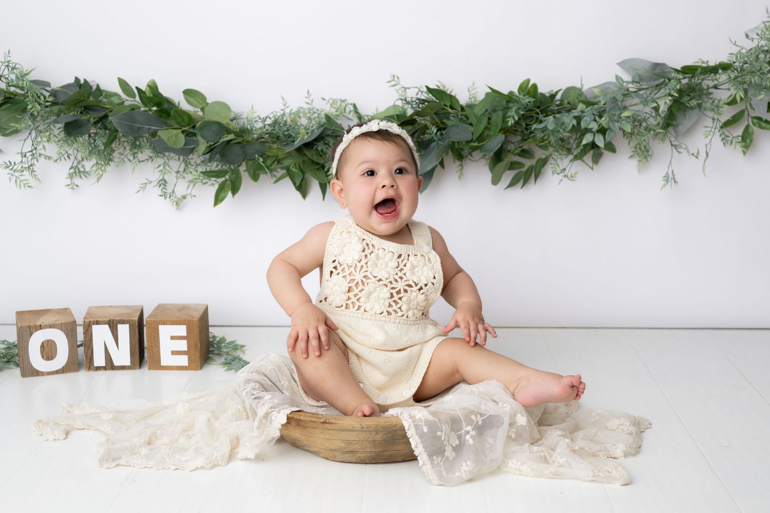 Baby in cream-colored outfit sits in wooden bowl against white backdrop with greenery garland decoration.