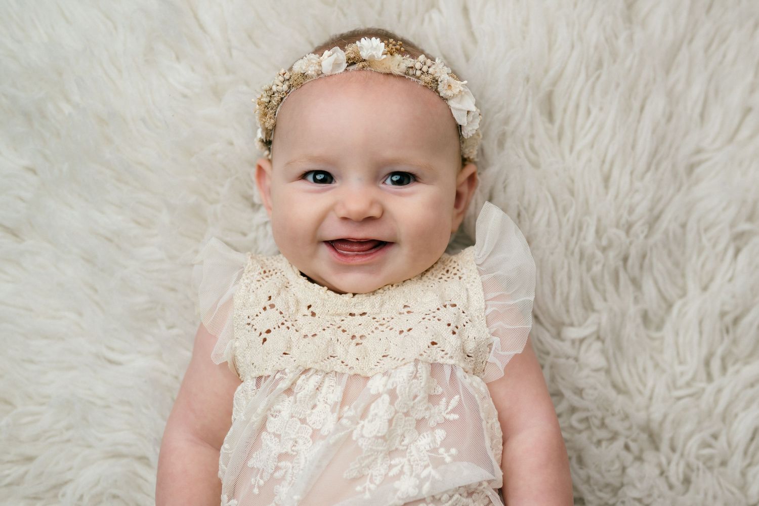 A baby wearing a delicate white lace dress and floral headband poses on a soft white fur backdrop.