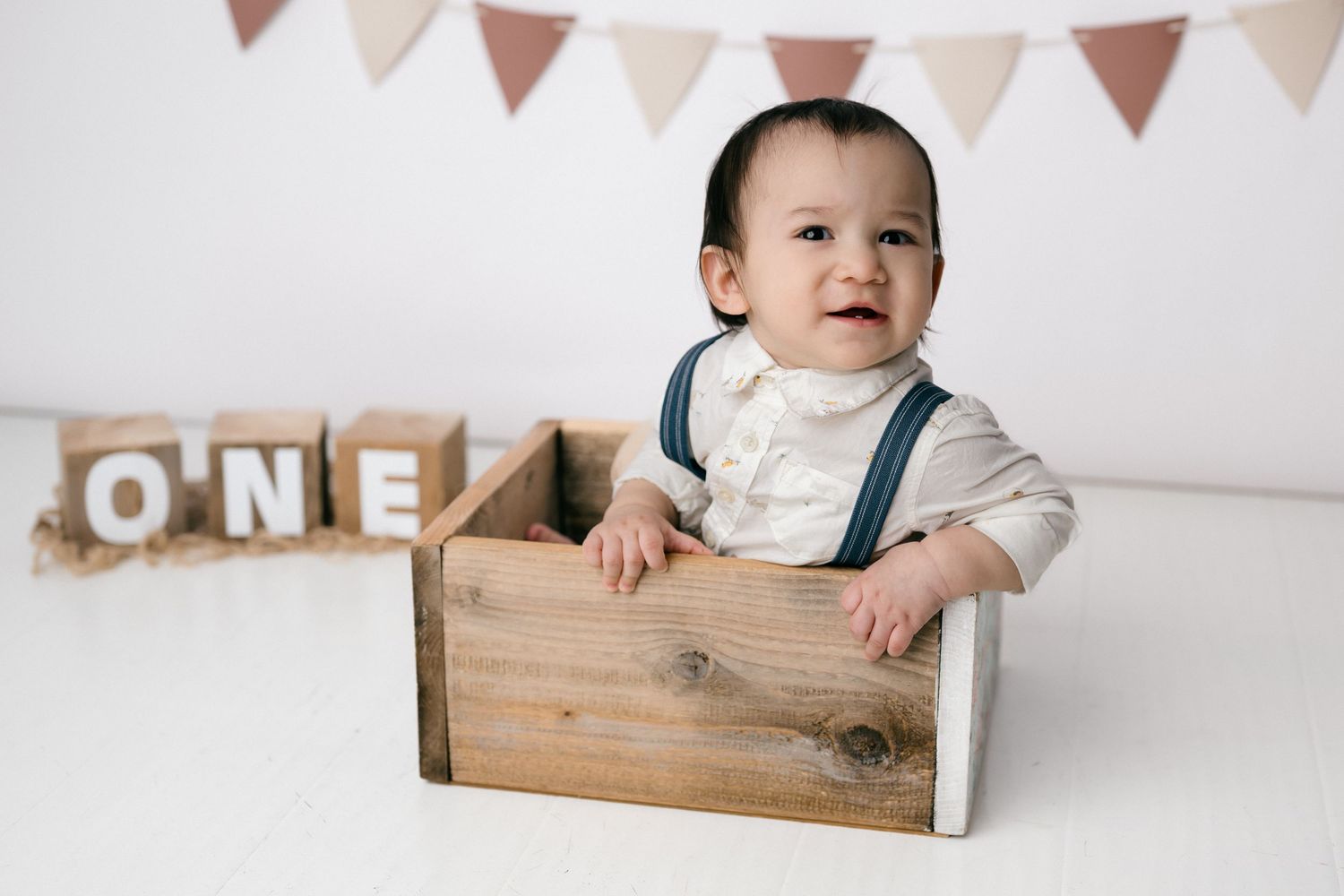 Baby boy in suspenders sits in a wooden crate beside ONE blocks at his first birthday photo shoot.