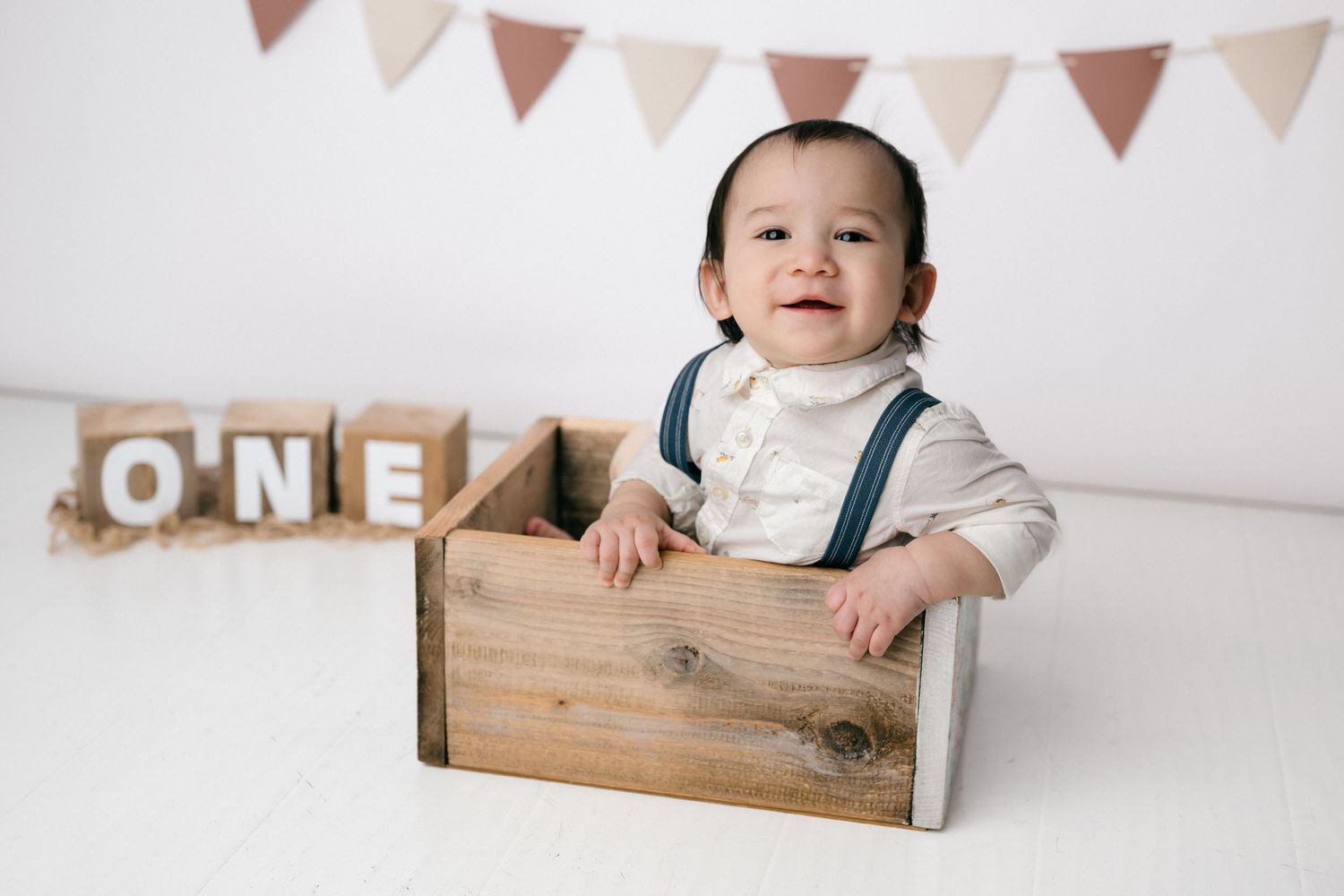 Smiling baby sitting in a wooden crate with ONE blocks beside them, celebrating their first birthday.
