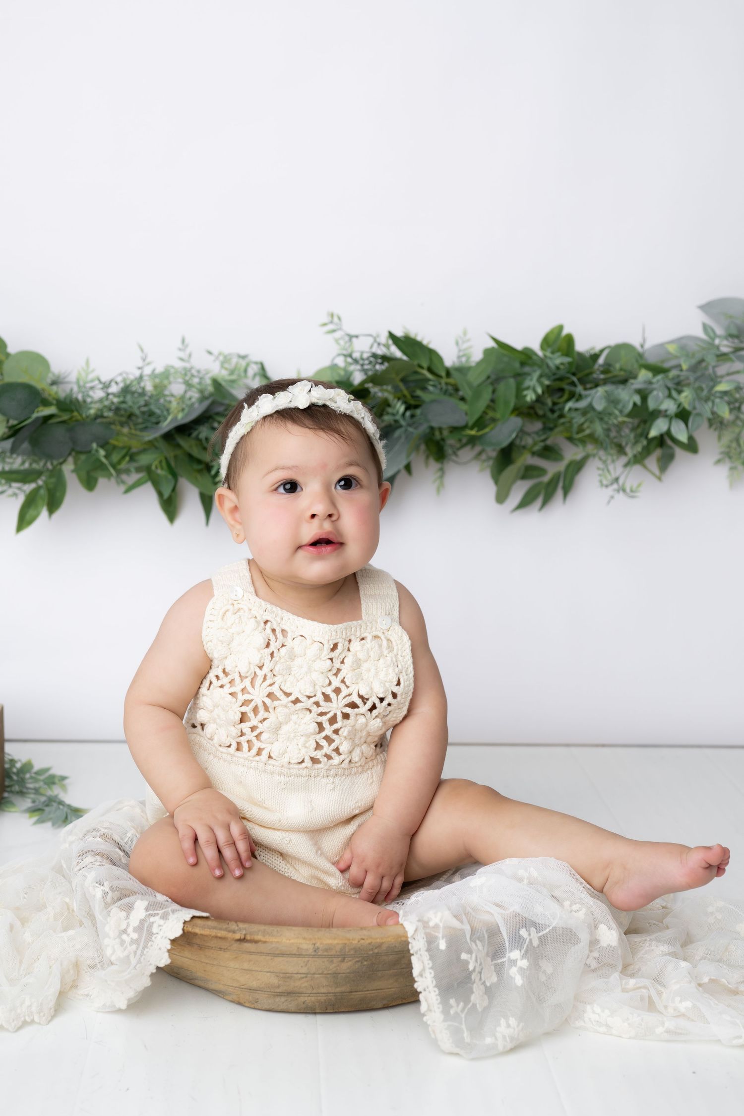 A baby wearing a cream colored eyelet dress sits in a wooden bowl with greenery and lace decorations.