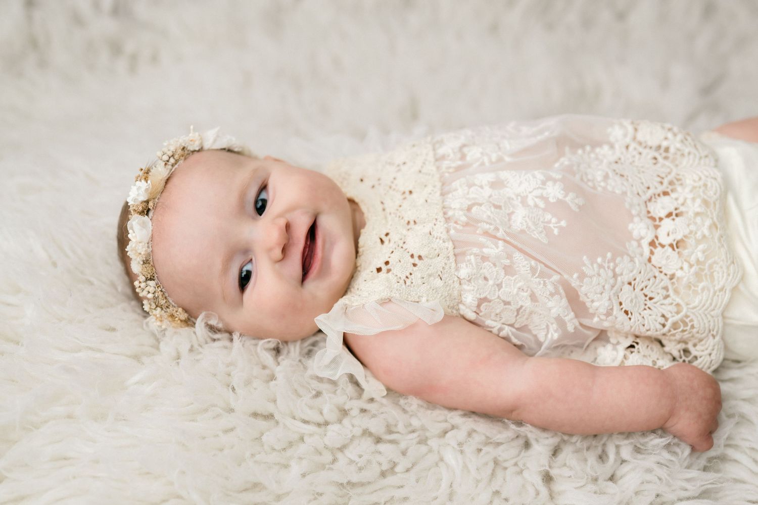 A baby wearing a white lace dress lies on a soft white blanket in a series of connected photos.