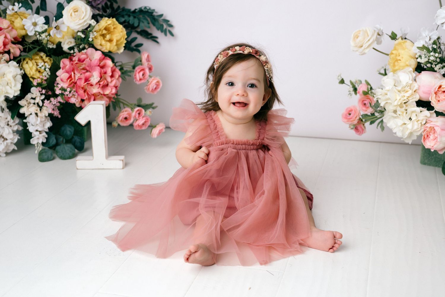 Baby girl in pink tulle dress sits smiling among flowers beside number 1, wearing floral headband.