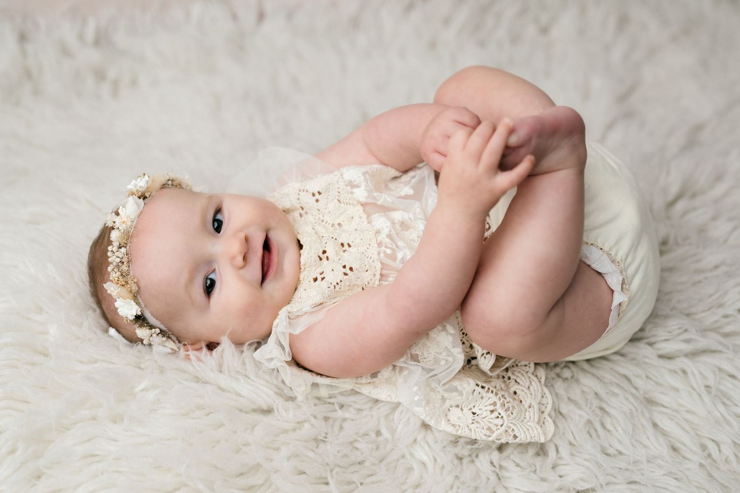 A baby wearing a delicate lace outfit lies on soft white fabric in a series of gentle poses.