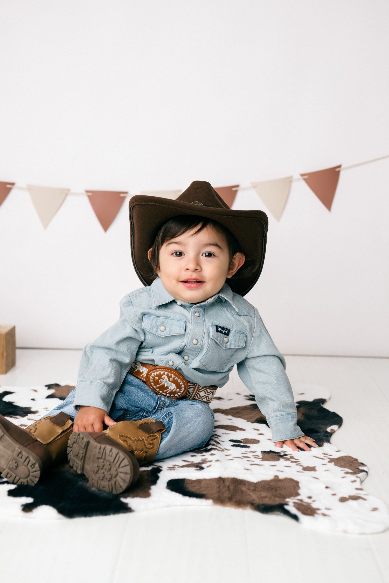 A small child in denim and cowboy boots poses on a white backdrop with brown cowhide print and festive bunting.