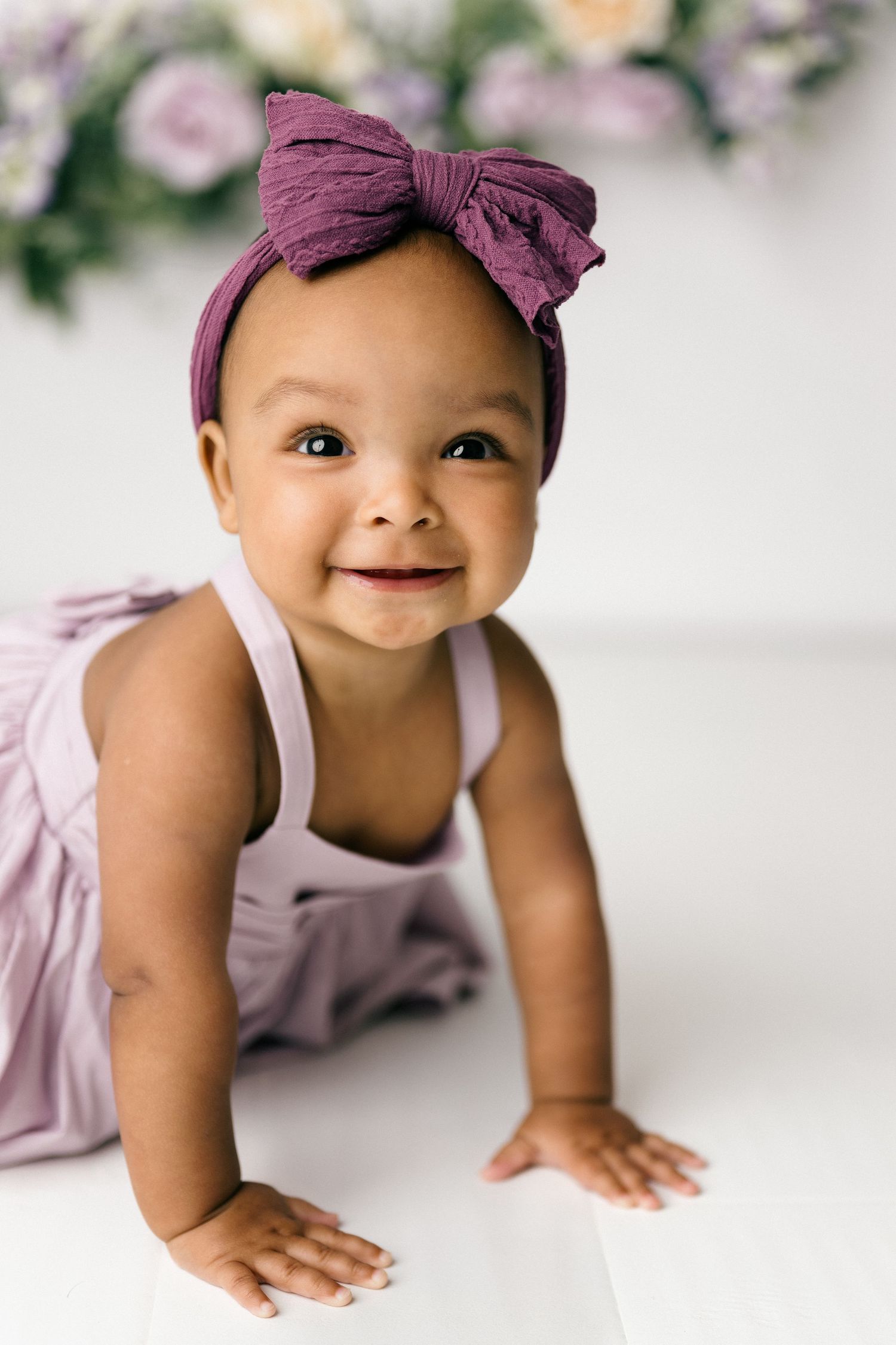 Baby wearing purple headbow and pink dress crawls on white surface with floral backdrop.