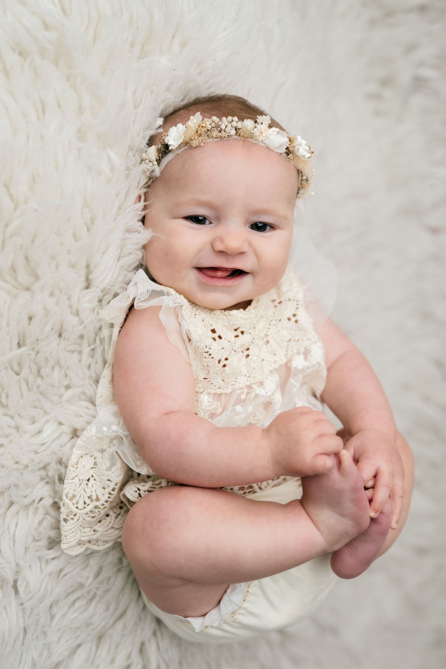 Baby wearing white lace romper and floral headband lies on soft white fur blanket in professional studio portrait.