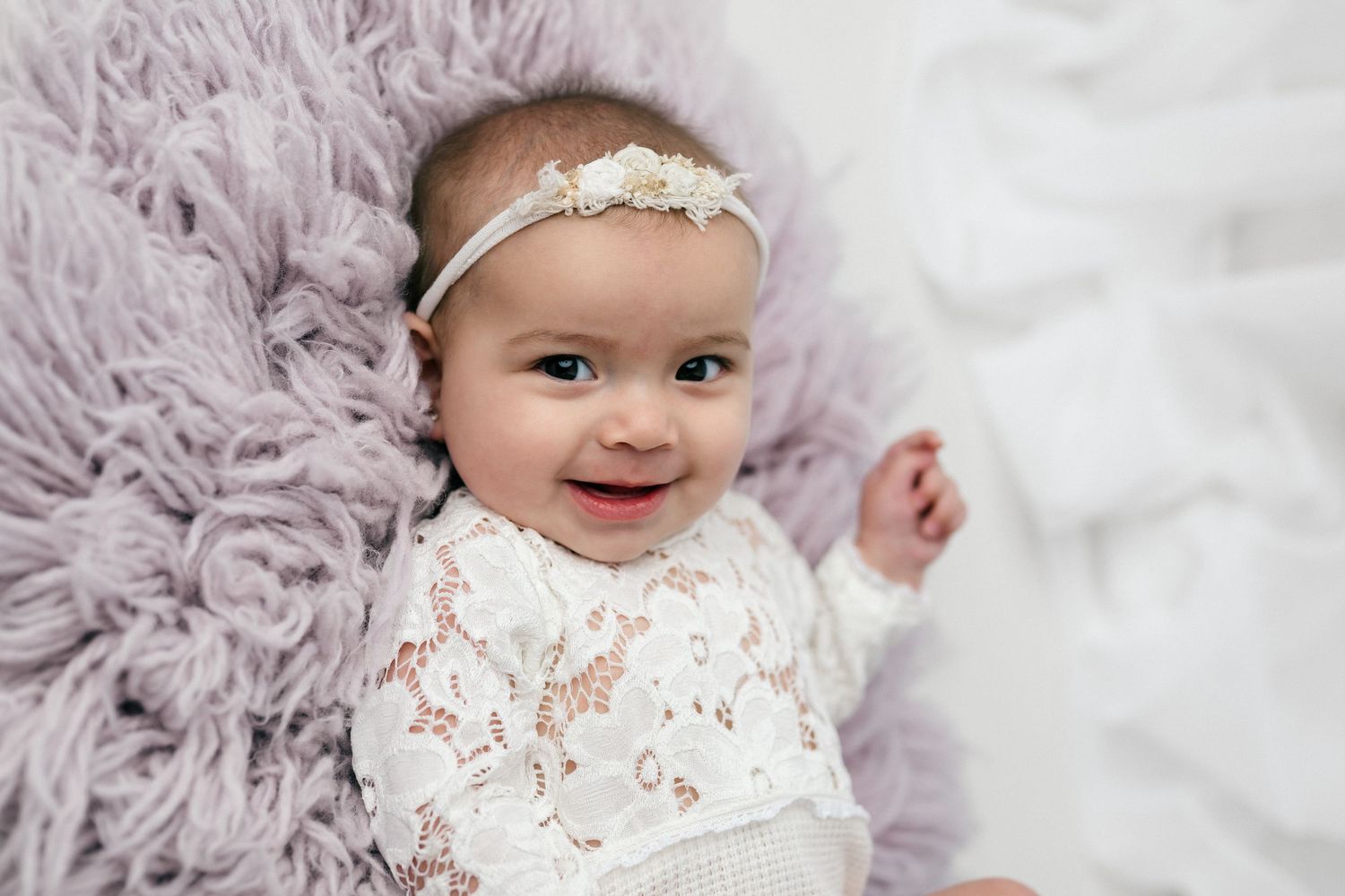 Smiling baby girl in white lace outfit and floral headband lying on a soft lavender fur blanket.