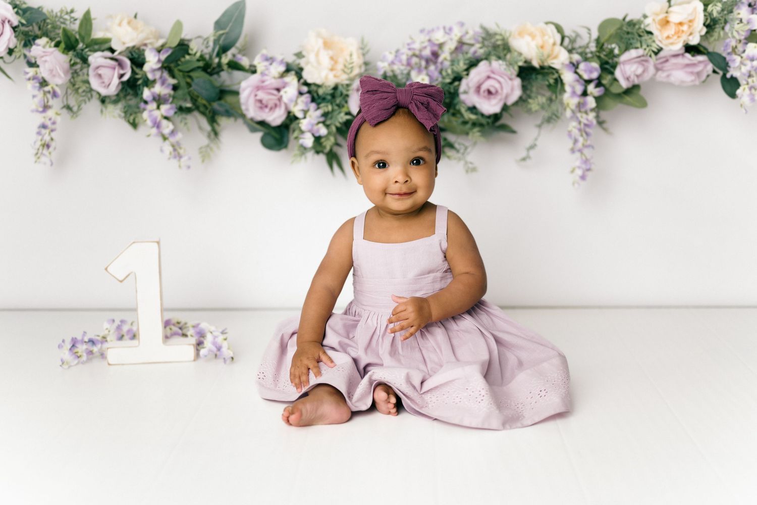 Baby in pink dress sits near floral arch with number 1 decoration against white background for first birthday photo.