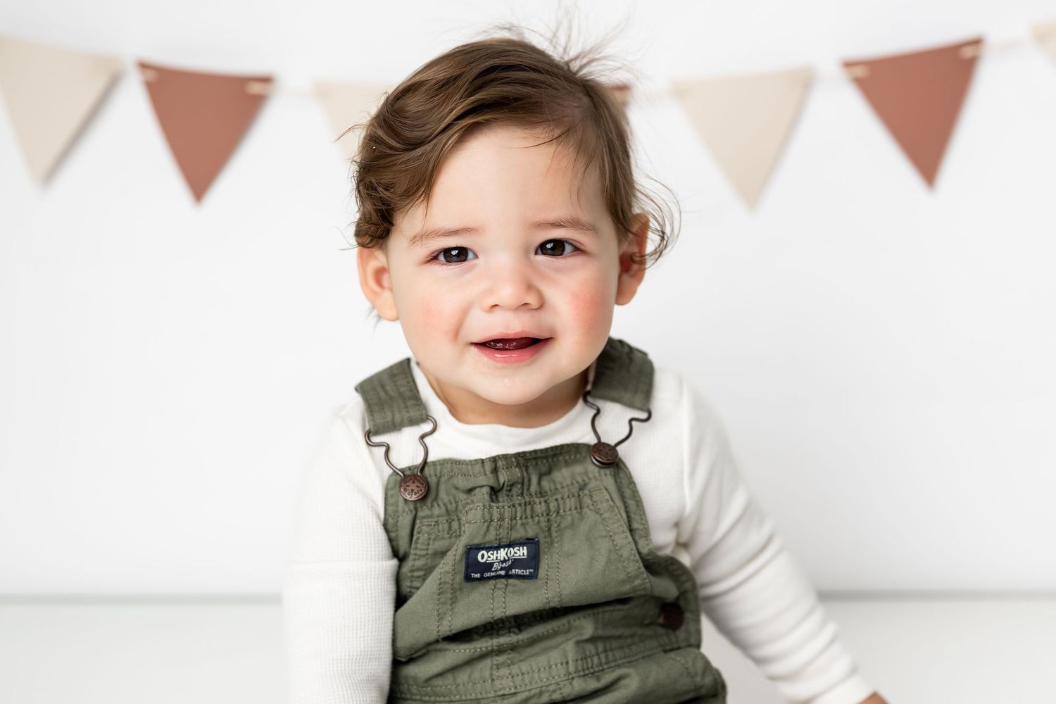 Smiling toddler wearing olive green overalls and white long sleeve shirt sits in front of triangle banner decorations.