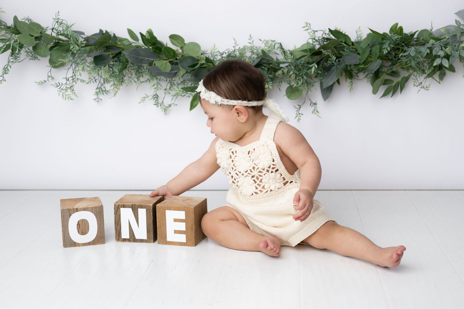 A baby in white sits on a white backdrop with greenery garland decorations for a first birthday photoshoot.