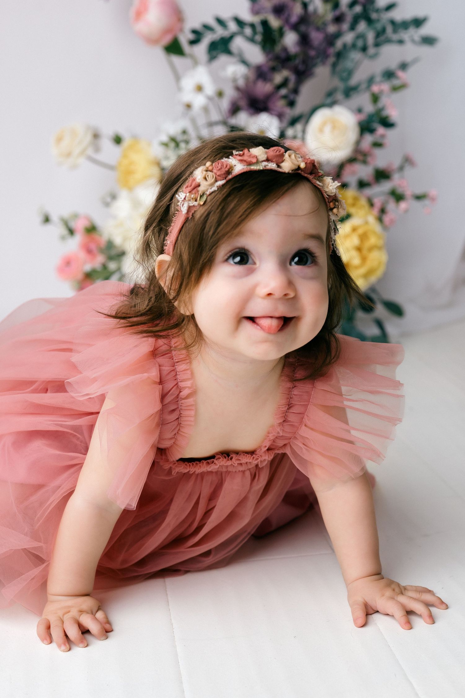 Adorable baby girl in pink tulle dress and floral headband crawling on white floor with colorful flowers behind her.