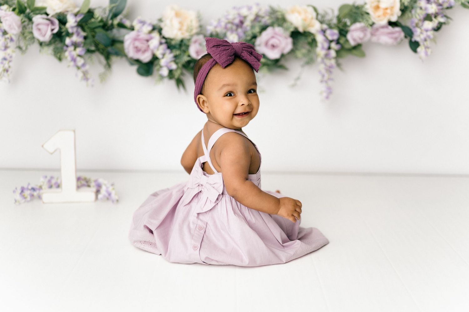 A baby in a lavender dress and purple bow sits against a white backdrop with floral garland decorations.