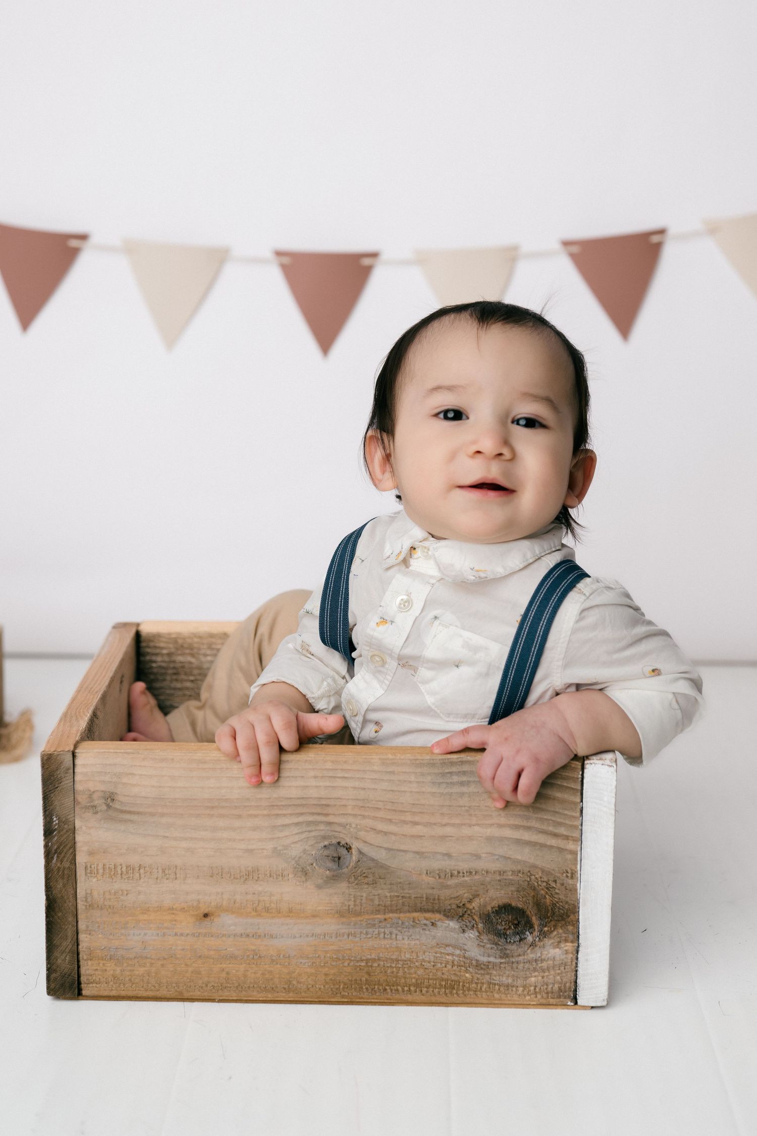 Adorable baby boy in white shirt and suspenders sitting inside a rustic wooden crate with festive bunting backdrop.