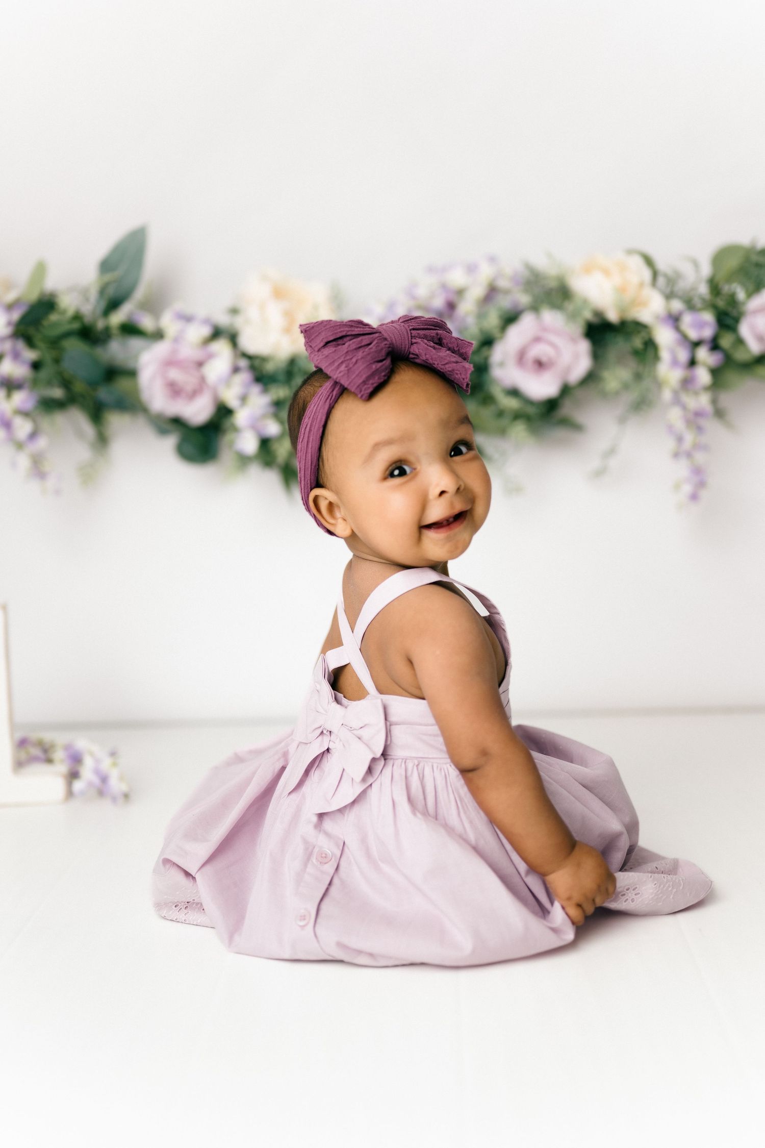 Happy baby wearing a purple dress and bow sits in front of a floral backdrop during a portrait session.