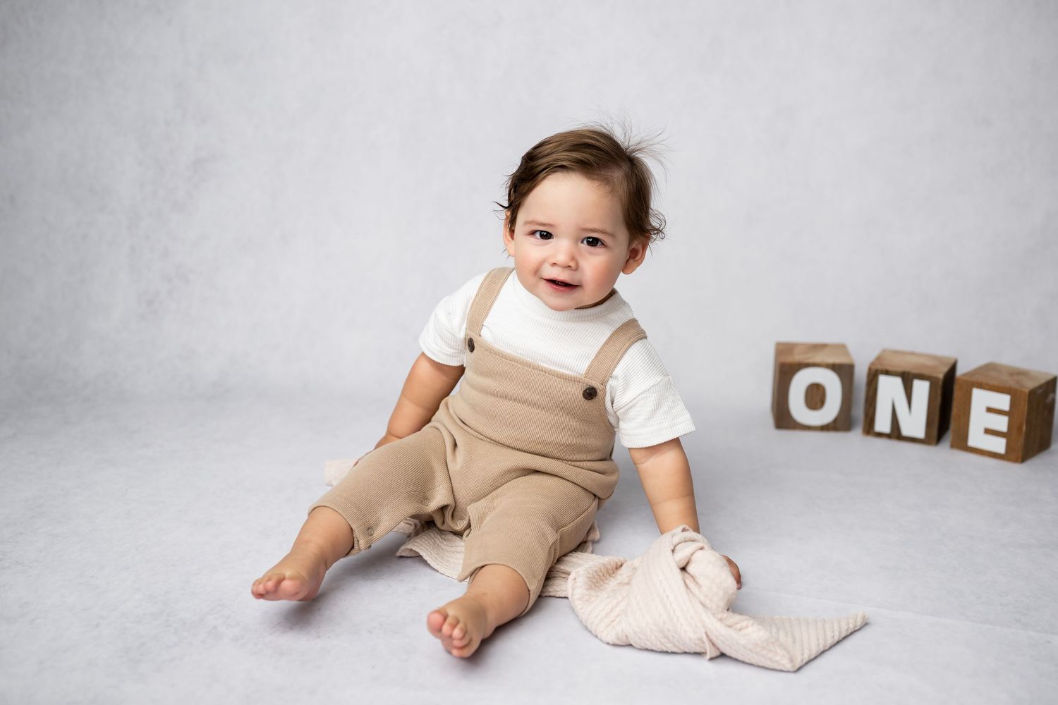 A toddler in tan overalls sits on gray background next to wooden blocks spelling ONE and plush toy.