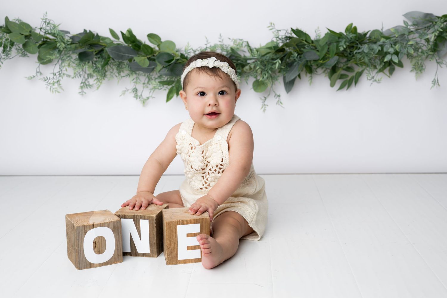 A baby wearing a white outfit sits on a white backdrop with green foliage garland and wooden letter decorations.