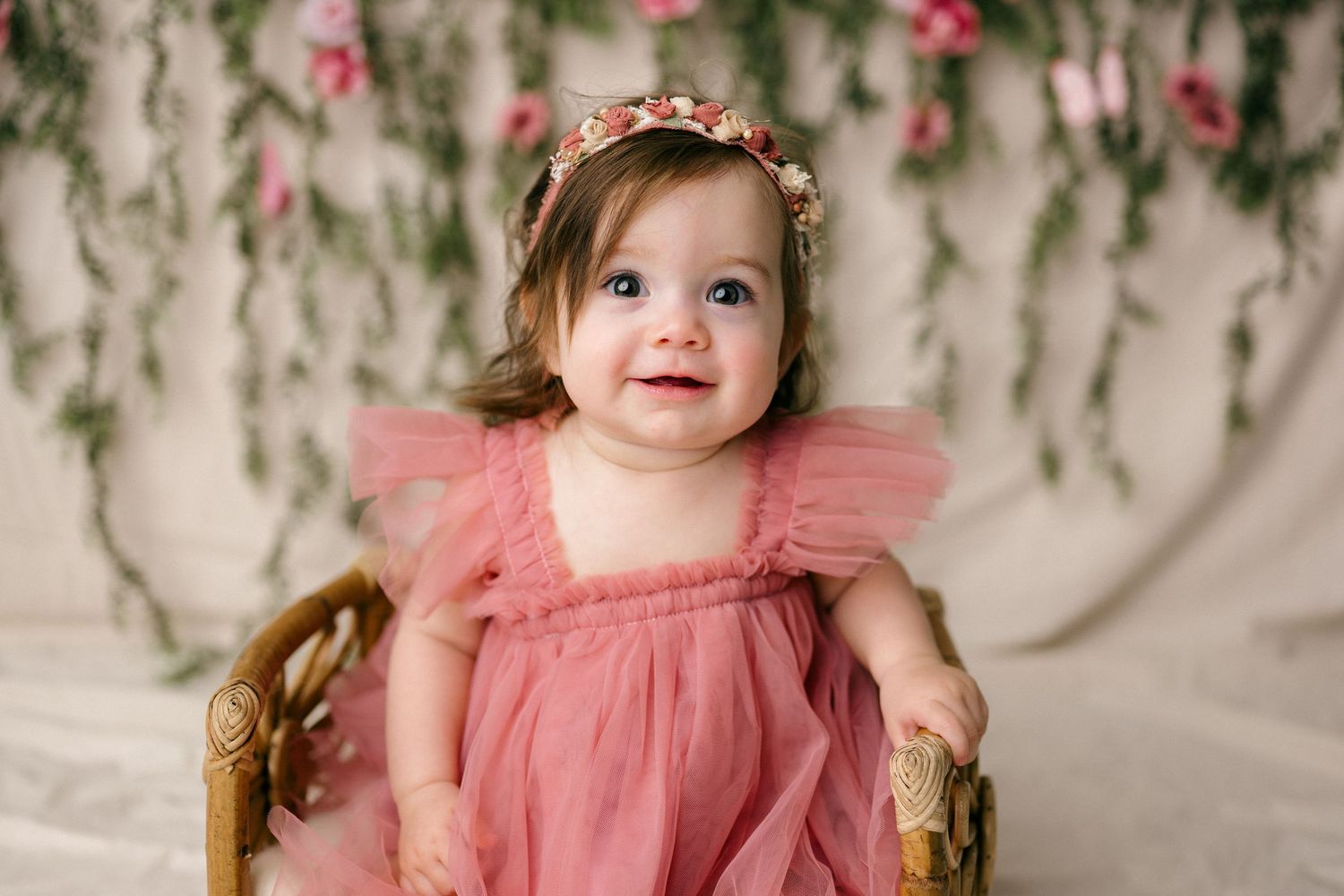 Smiling baby girl in pink tulle dress and floral headband sits in wicker chair with floral backdrop.