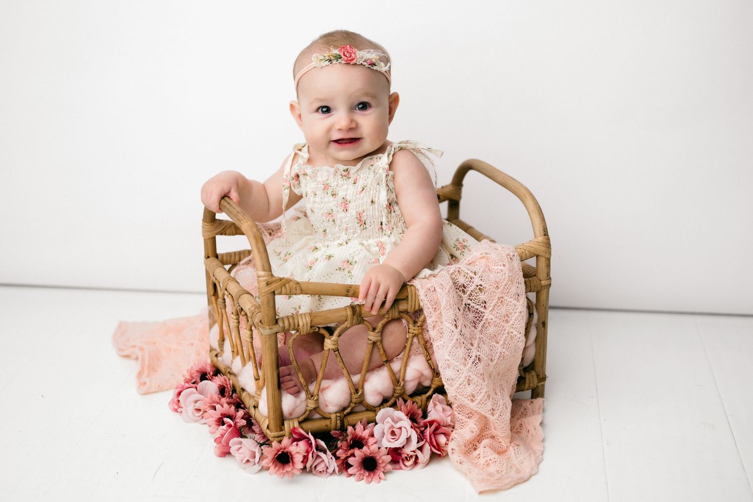Baby in floral dress sits in wicker basket surrounded by pink roses and blush knit blanket against white backdrop.