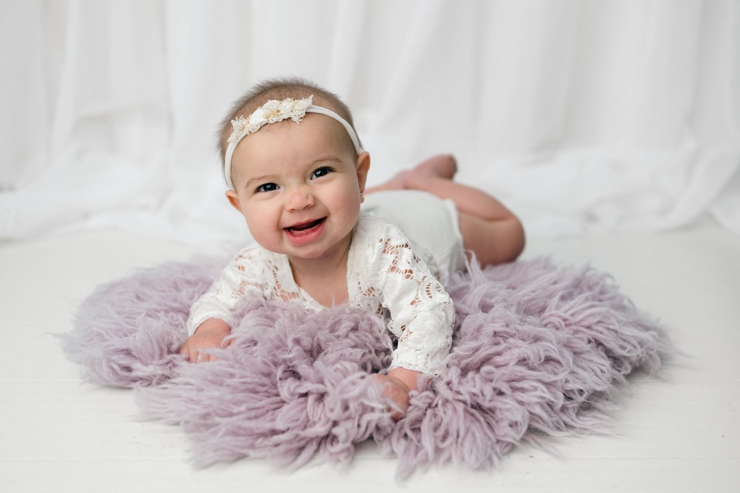 Smiling baby girl in white lace outfit lying on lavender fluffy rug with floral headband.