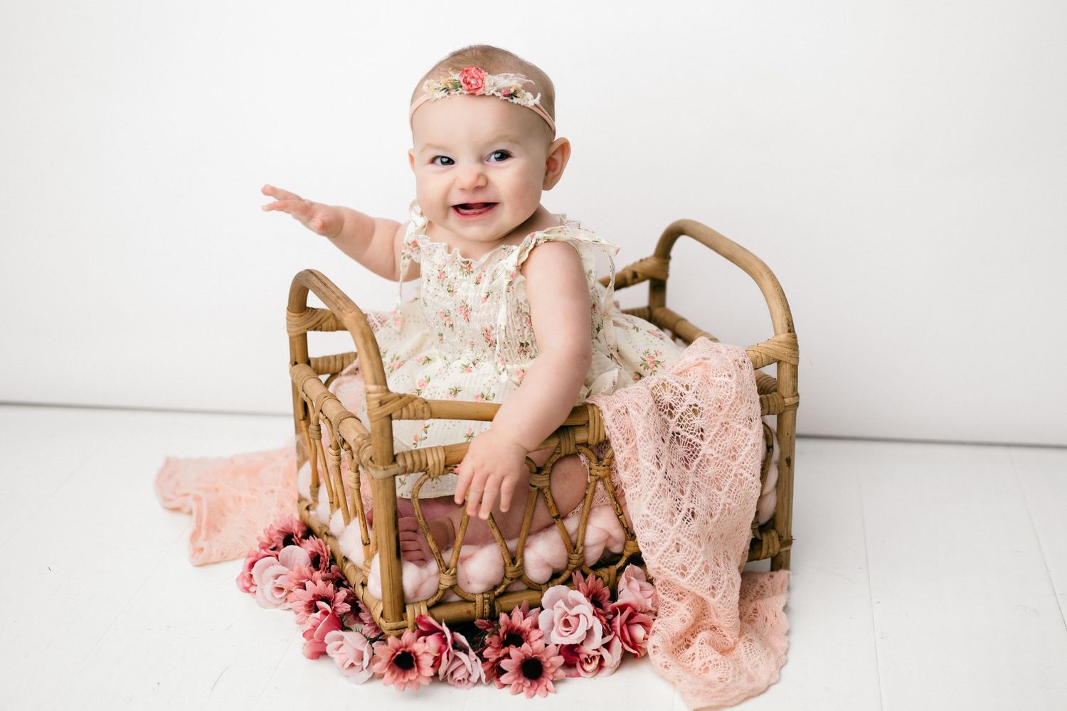 Baby wearing pink lace romper sits in wooden chair surrounded by pink flowers during professional photo session.