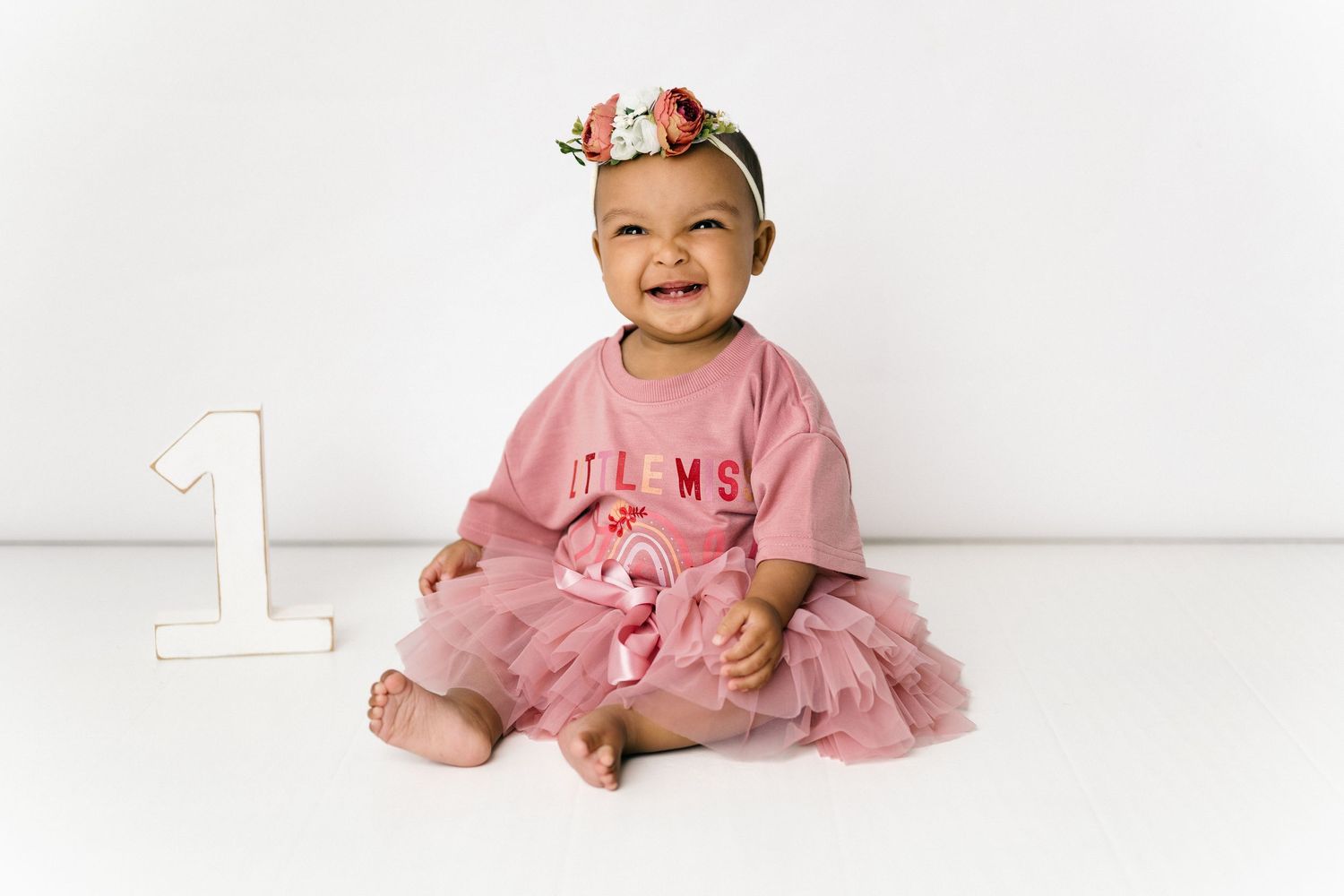 A baby wearing a pink tulle dress and floral headband sits next to a large number one on a white backdrop.