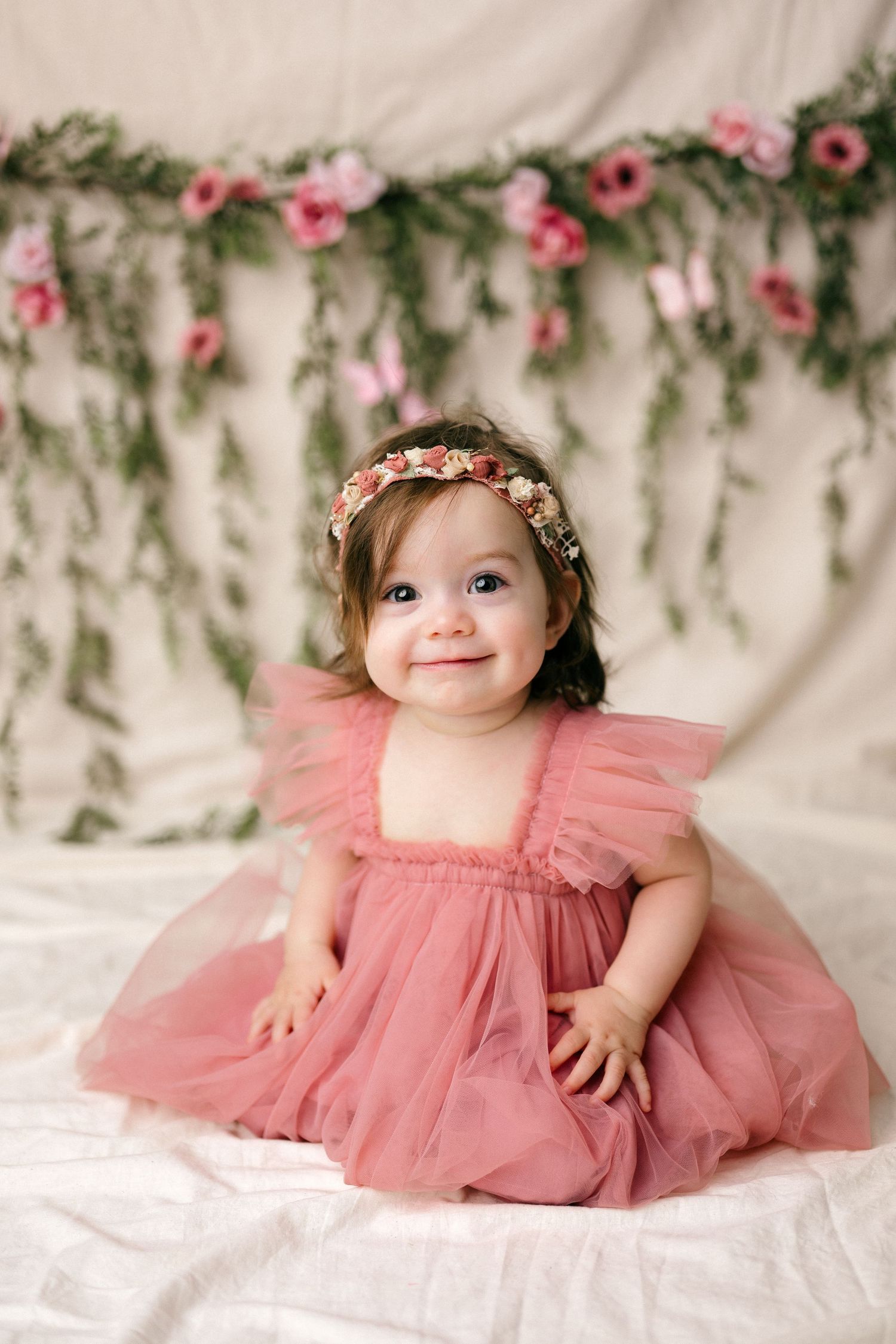 Smiling baby girl in pink tulle dress and floral headband sitting before a floral garland backdrop.