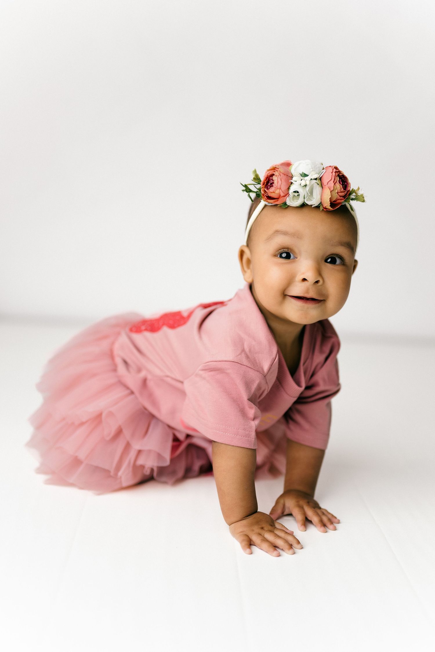 Baby wearing pink tulle dress and floral crown crawls on white studio background.