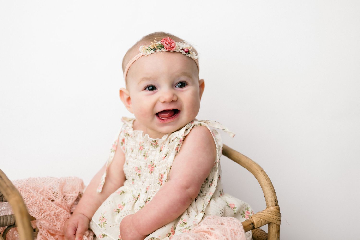 Baby wearing a cream lace dress and floral headband smiles while sitting in a wooden chair.