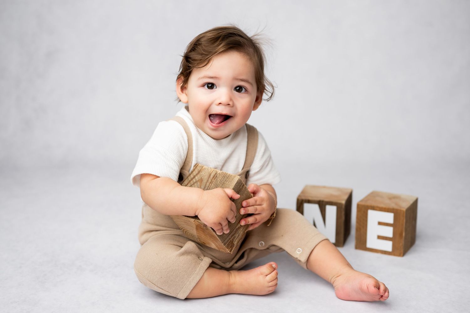 A young child in beige overalls sits on a light floor next to wooden alphabet blocks displaying 'E'.