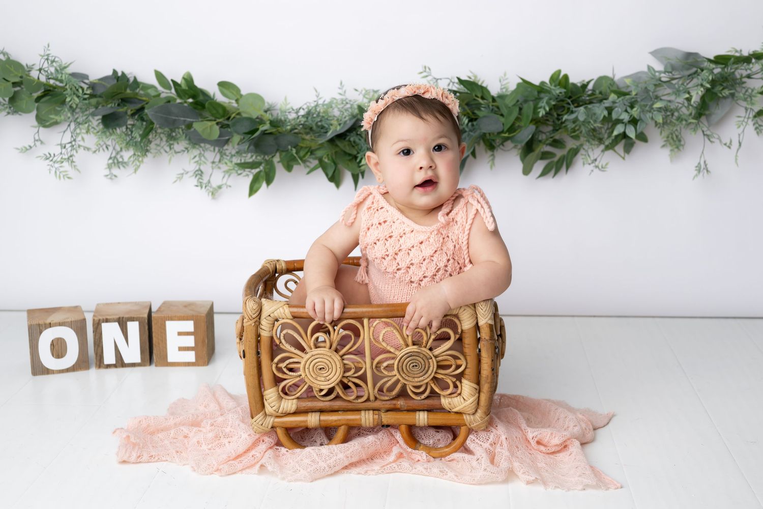 A baby in a pink dress sits in an ornate gold chair with greenery garland decorations in the background.