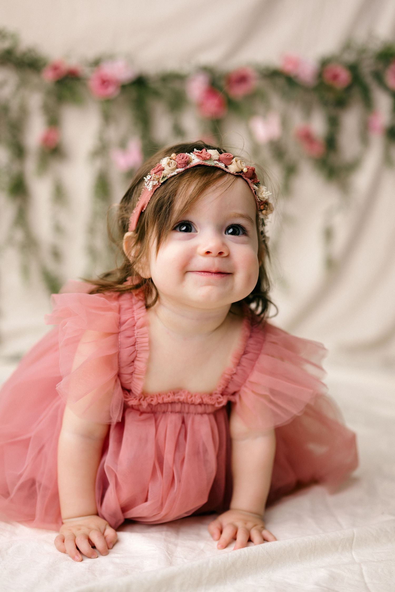 Adorable baby girl in pink tulle dress and floral crown smiles while posing in a dreamy floral studio setting.