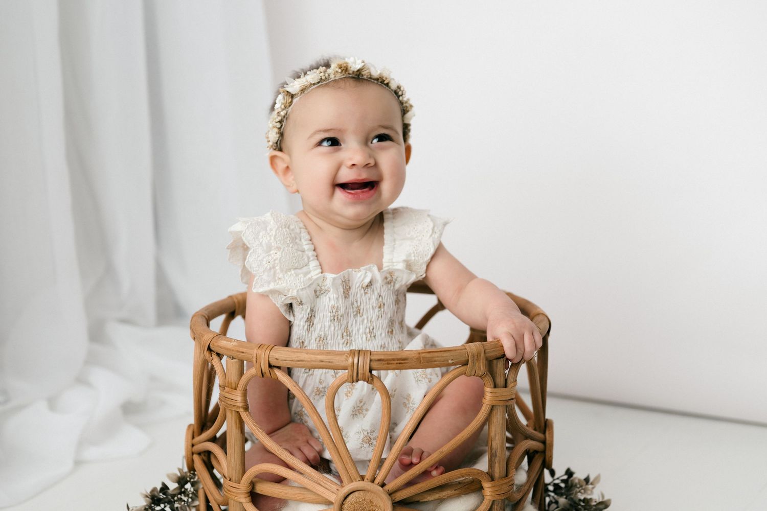 Smiling baby girl in white lace romper sitting in a wicker basket with floral headband in studio.