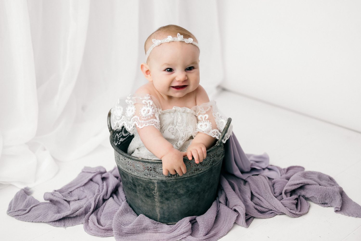 A baby sits in a dark green bucket surrounded by flowing purple fabric in a bright white studio setting.