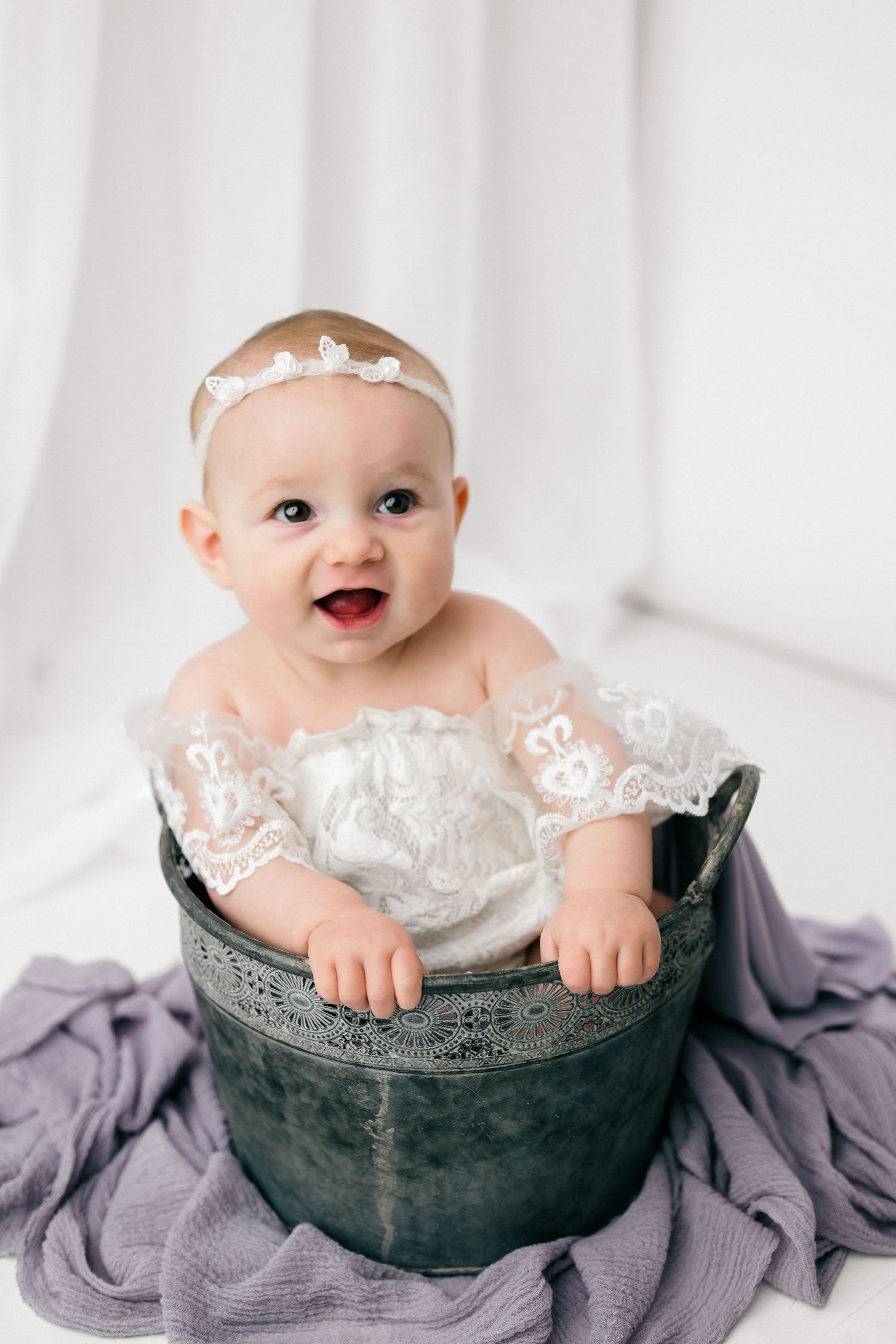 A smiling baby wearing a white lace headband sits in a vintage metal basin on purple fabric.