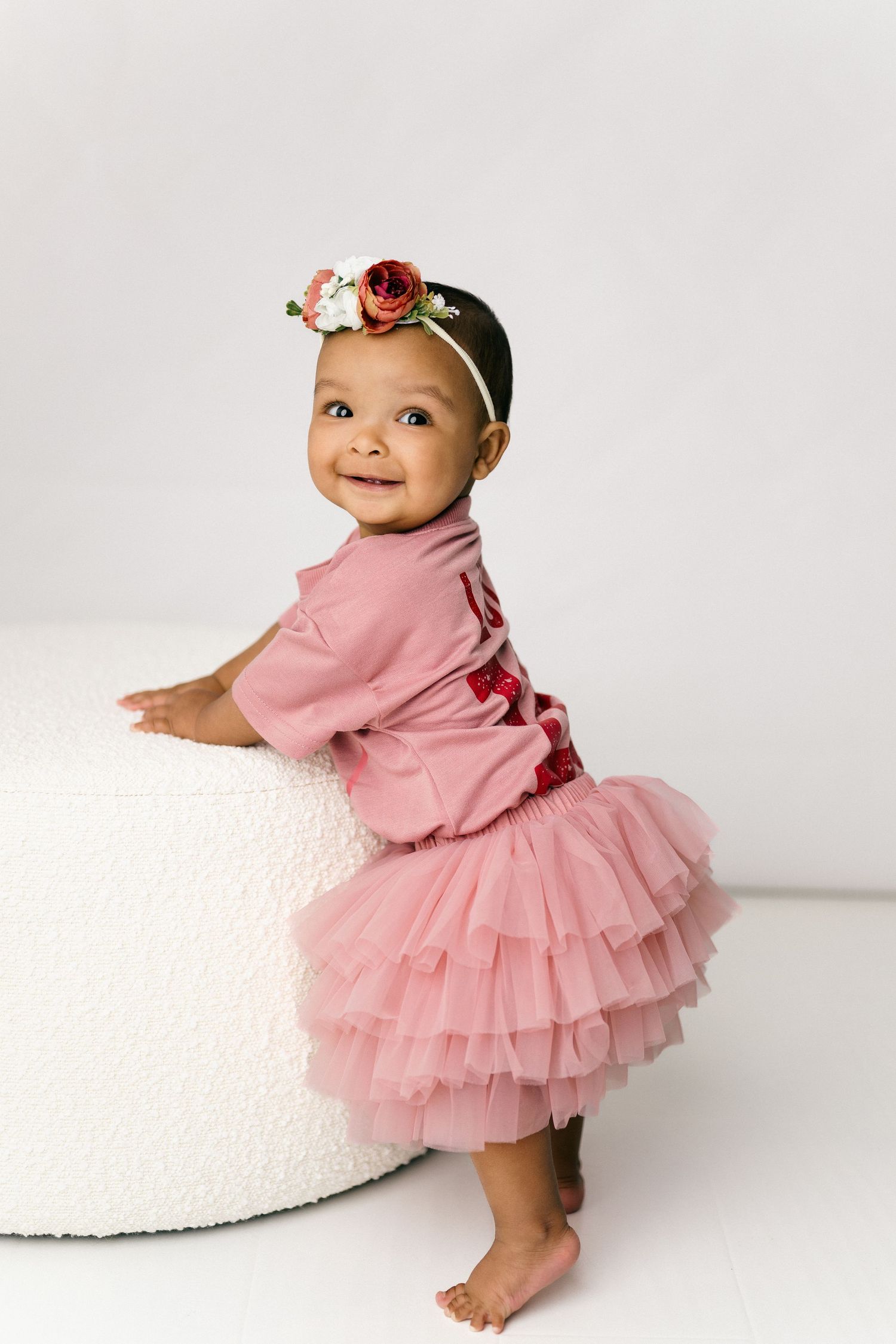 Baby wearing pink tutu dress and floral headband poses on white curved ottoman against light background.