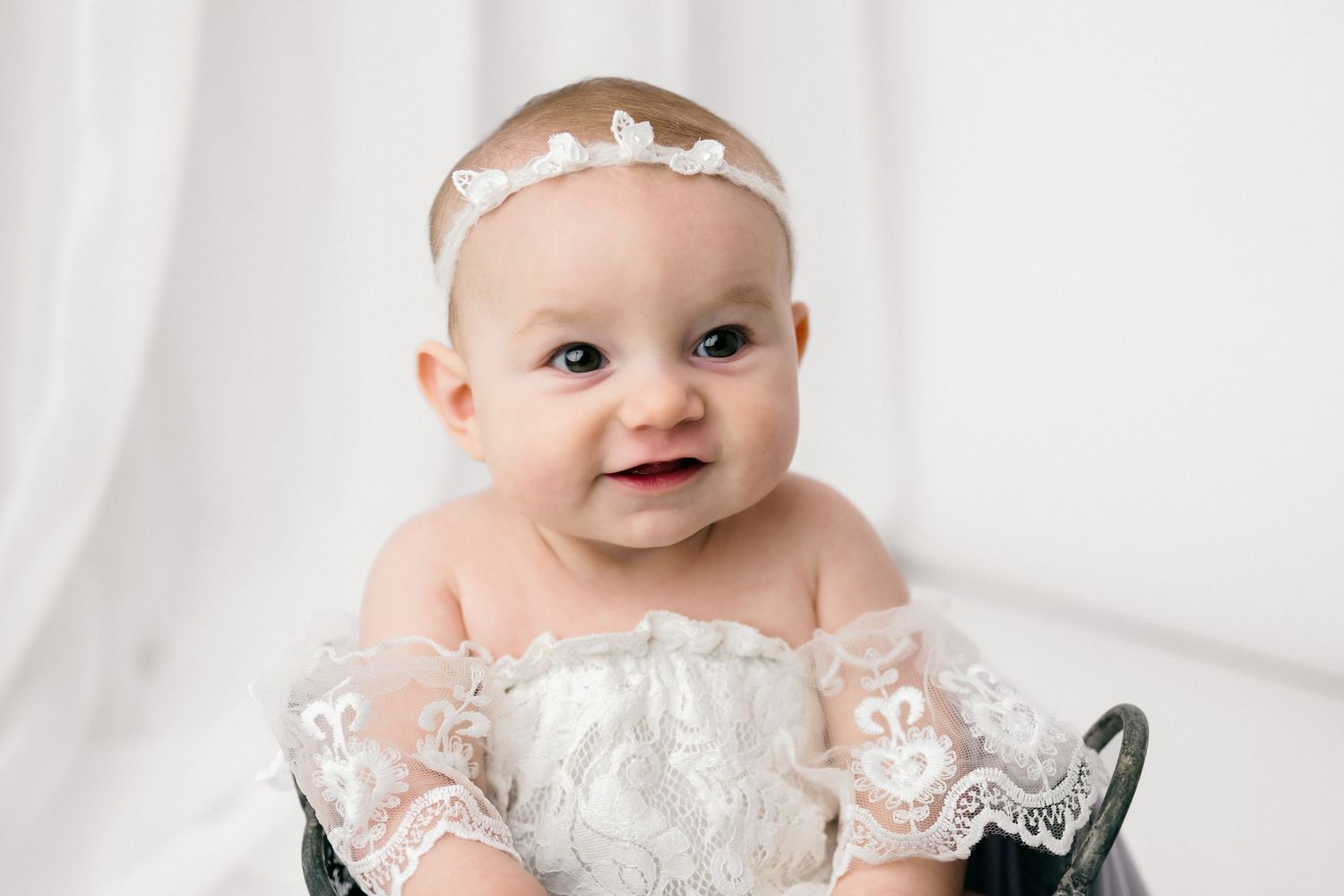 Baby wearing white lace dress smiles while sitting against white backdrop in studio portrait setting.