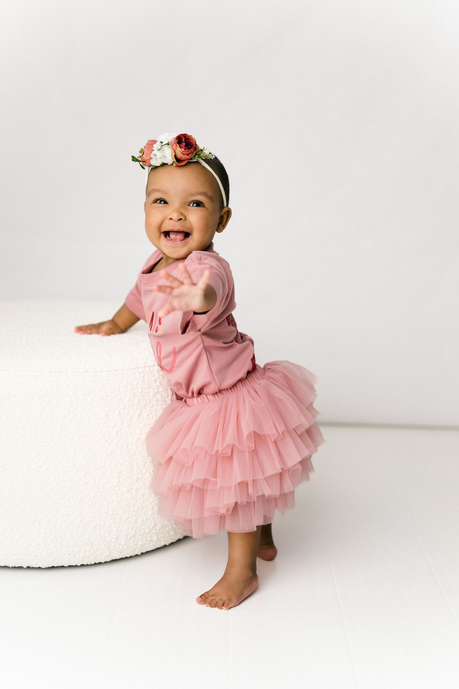 Smiling baby in pink tulle dress and floral headband stands barefoot against white background.