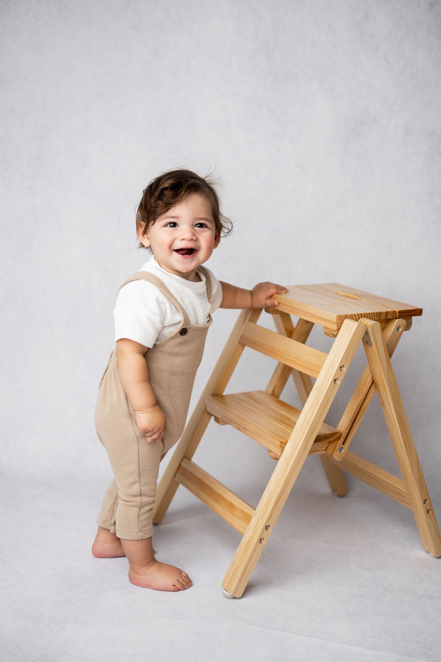 Adorable toddler in beige overalls smiles while standing next to a wooden step stool against a gray backdrop.