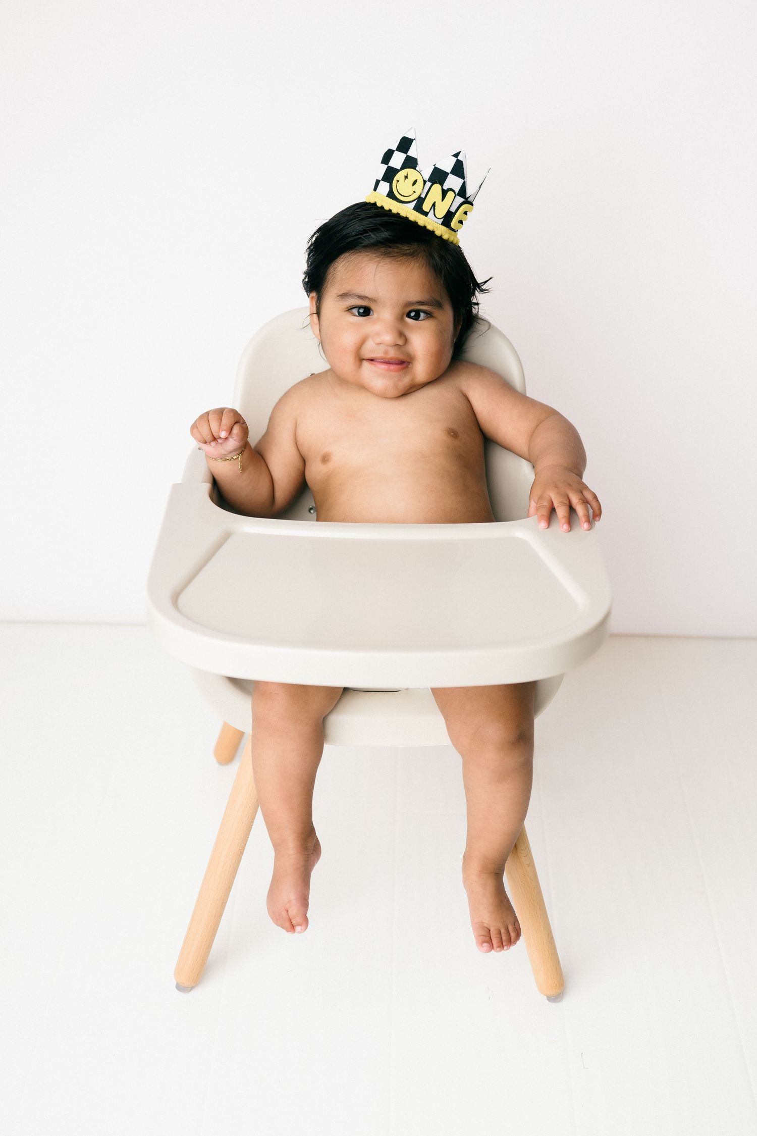 Baby wearing a tiny crown sits in white high chair against plain white background, smiling at camera.